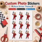 A sheet of custom photo stickers with a race driver theme, featuring cartoon toddlers in red racing suits holding checkered flags. The sheet is displayed on a light wood desk surrounded by craft supplies like scissors, tape, ribbon, and twine.