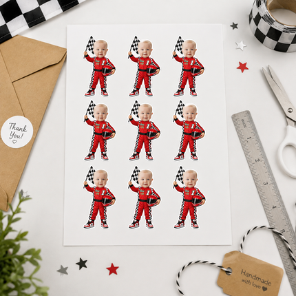 A sheet of nine identical cartoon baby racecar stickers in red racing suits with checkered flags, displayed on a white desk with a kraft envelope, scissors, ruler, tape, and star decorations.