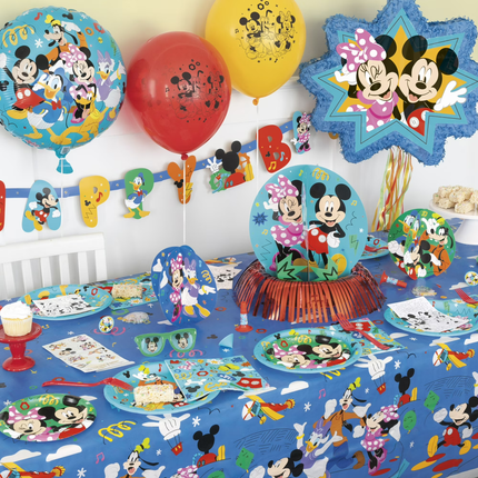 A Mickey Mouse and Minnie Mouse themed party table setting featuring a blue printed tablecloth, matching plates, napkins, balloons, and decorative banners on a white wall.