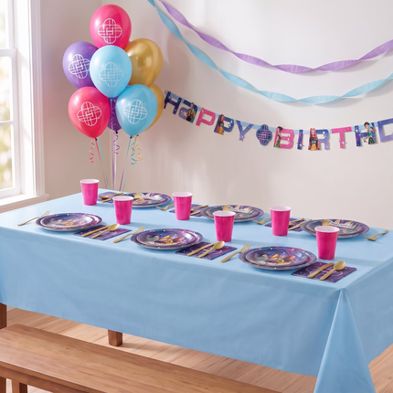 A light blue party tablecloth set on a wooden table, styled with matching blue plates, pink cups, gold cutlery, and purple napkins. The scene includes a balloon bouquet, a KPop Demon Hunters Happy Birthday Banner, and paper streamers hanging on the wall.