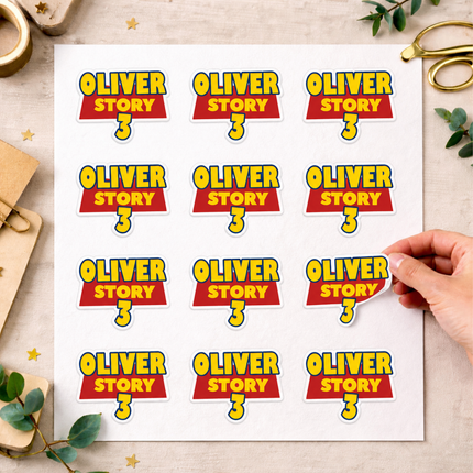 A sheet of die-cut Toy Story stickers featuring the 'Oliver Story 3' logo, held by a hand on a styled flat-lay desk background with gold scissors, eucalyptus leaves, and a gold pen.